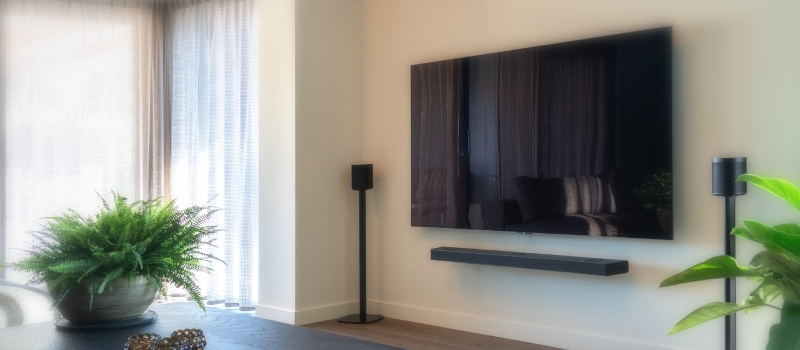 Wohnzimmer mit weißen Vorhängen links und einem Fernseher mit Soundbar und zwei separaten Lautsprechern auf Lautsprecherständern rechts an der Wand